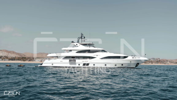 A luxury motor yacht anchored in a turquoise bay with the iconic whitewashed buildings and windmills of Mykonos, Greece, in the background. Representing Mykonos Yacht Rental and Luxury Boat Charter Greece.